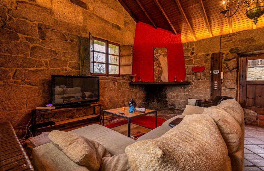 A rustic living area features exposed stone walls and wooden beams overhead. A comfortable sofa sits in front of a coffee table, while a television is positioned on a stand. The red accent wall adds warmth to the space, complemented by a fireplace at the far end.