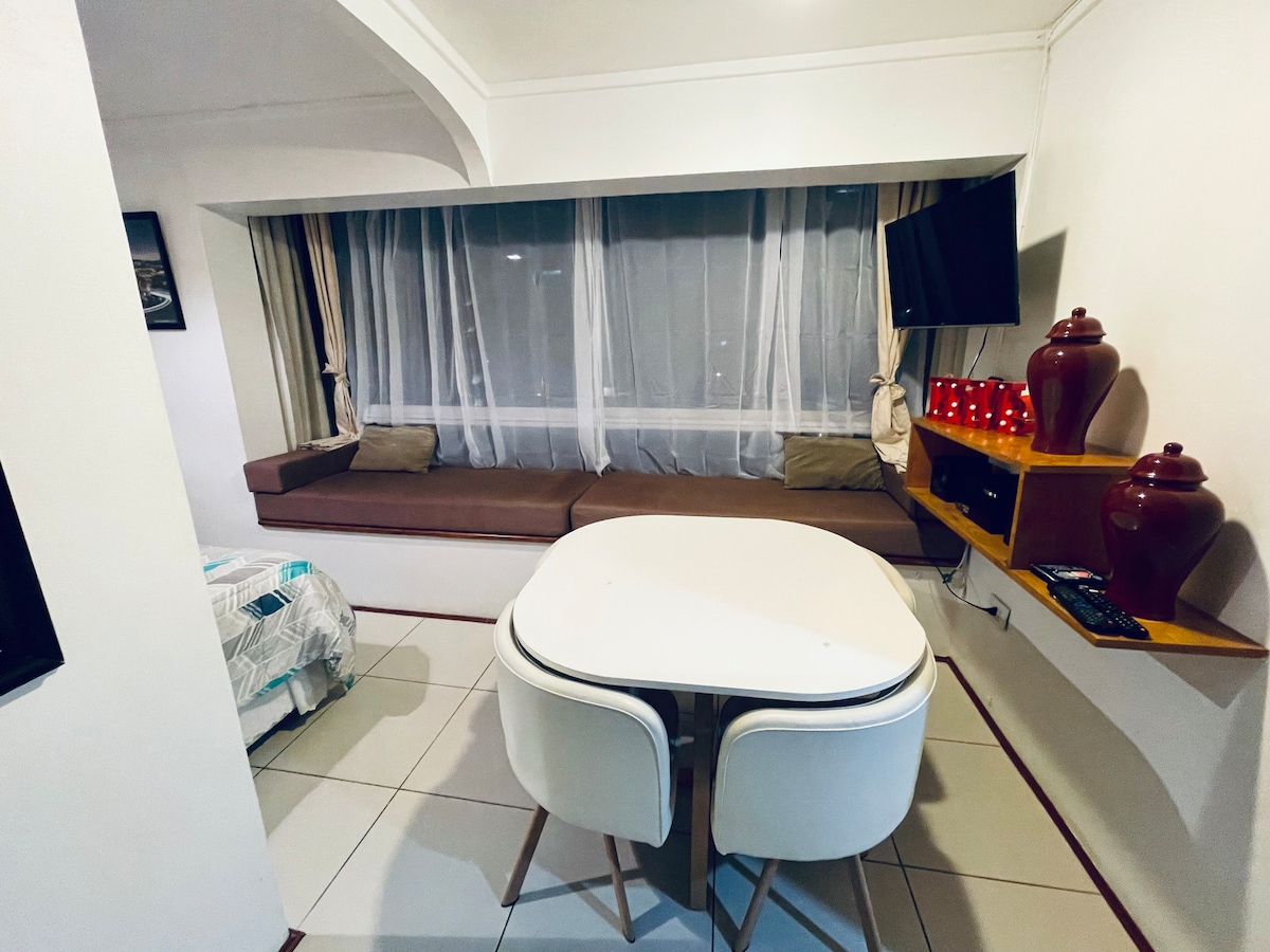 A well-lit living area features a comfortable brown seating nook beside a large window dressed with sheer curtains. A round dining table with light-colored chairs is positioned adjacent to a wall-mounted television. Decorative red vases are displayed on a mounted shelf, adding a touch of style.