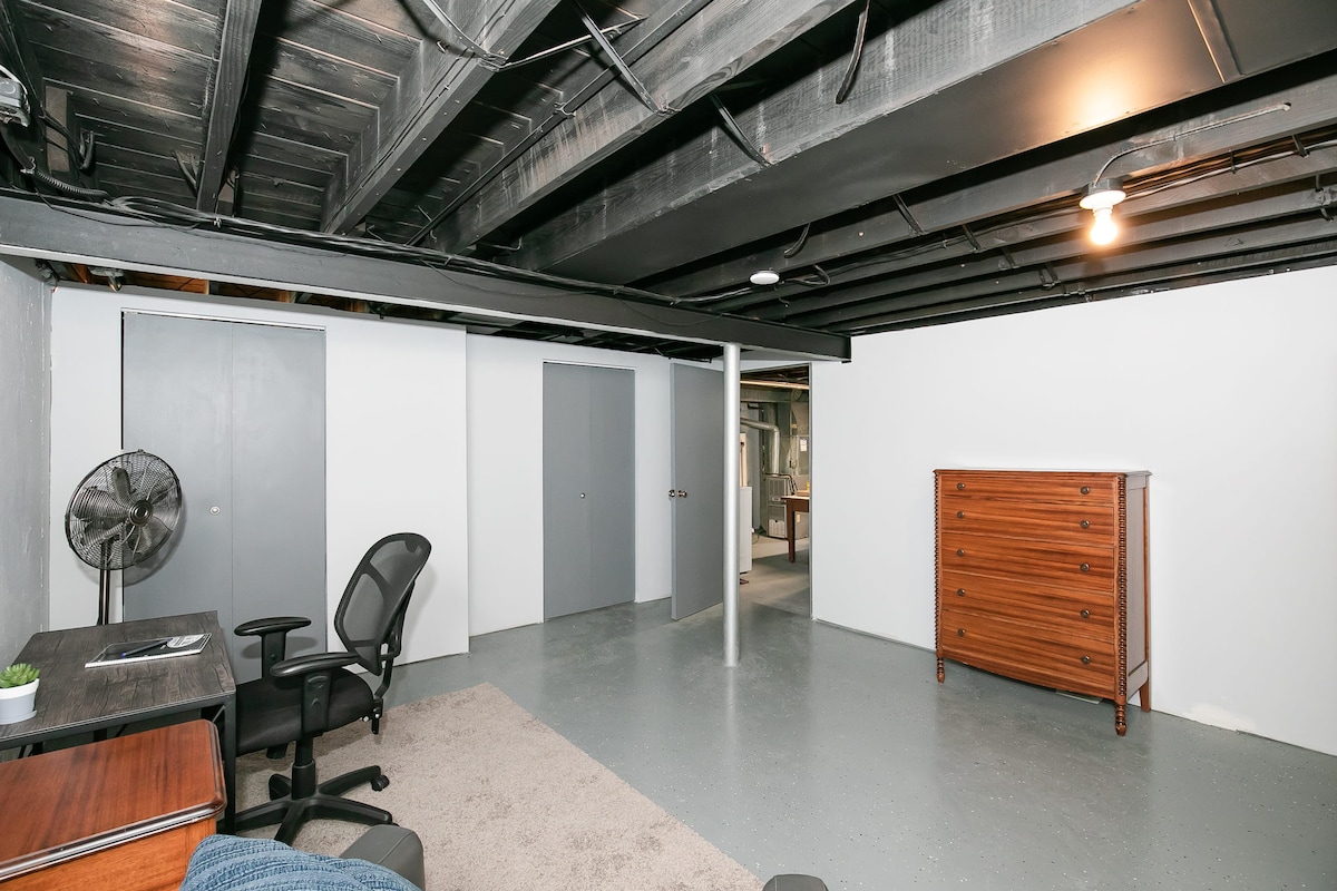 An open basement space is shown, featuring painted concrete floors and exposed black beams overhead. A small wooden dresser is positioned against one wall, while a desk and chair are located nearby. A fan rests on the floor, and neutral-colored walls create a bright ambiance.