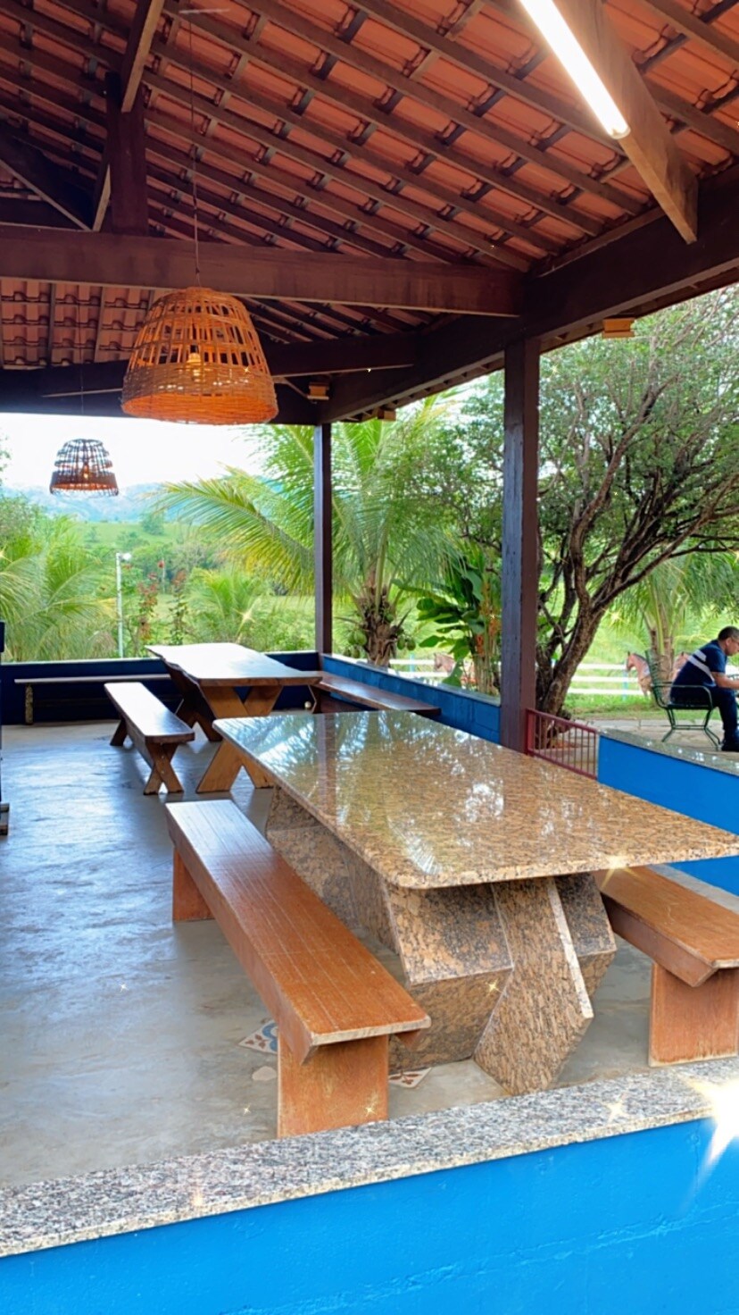 A covered outdoor dining area features a large granite table surrounded by wooden benches. The space is open to nature, showcasing green foliage in the background. Hanging light fixtures provide illumination, while the roof is supported by wooden beams, enhancing the rustic charm.
