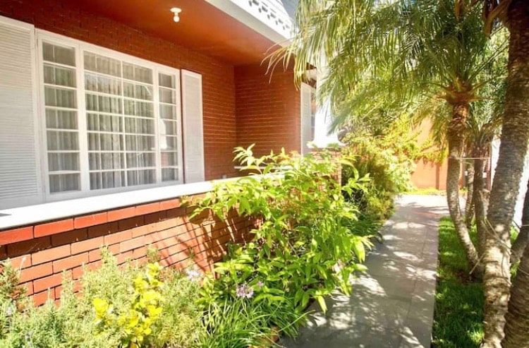 A welcoming exterior view showcases a pathway lined with lush greenery and colorful flowers. The brick facade is complemented by white-framed windows, allowing natural light to brighten the surrounding area.