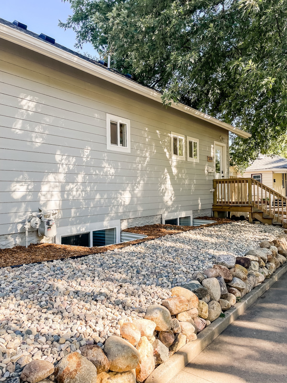 The exterior view showcases a light gray house with a wooden deck and steps leading to the entrance. A well-maintained path is lined with smooth stones and boulders, while several small windows are visible along the side of the house, surrounded by greenery.