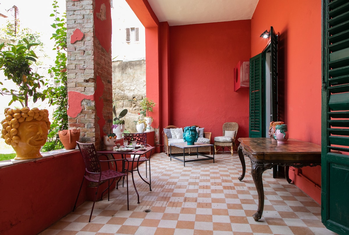 A charming patio area is featured with tiled flooring and red walls. A wooden table and several chairs surround a small seating arrangement, complemented by decorative pottery and plant arrangements. Natural light illuminates the space, creating an inviting atmosphere for relaxation.