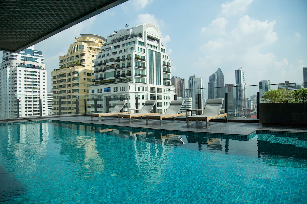 A rooftop pool is surrounded by sleek lounge chairs, offering a panoramic view of the city skyline. The pool's water glistens under the daylight, reflecting the modern high-rise buildings nearby.