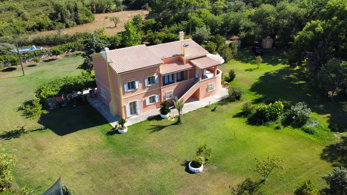 An aerial view showcases a spacious garden house surrounded by lush greenery, including various trees and plants. The property features a well-maintained lawn and is complemented by clear blue skies. The house is painted in warm tones, with visible balconies and a tiled roof.