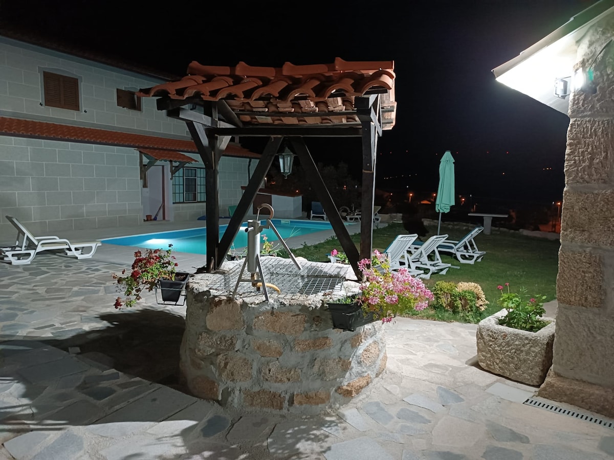 A stone well with a wooden structure is surrounded by blooming flowers and a paved area. In the background, a pool is illuminated against the night sky, and several lounge chairs are positioned nearby, creating a peaceful outdoor space.