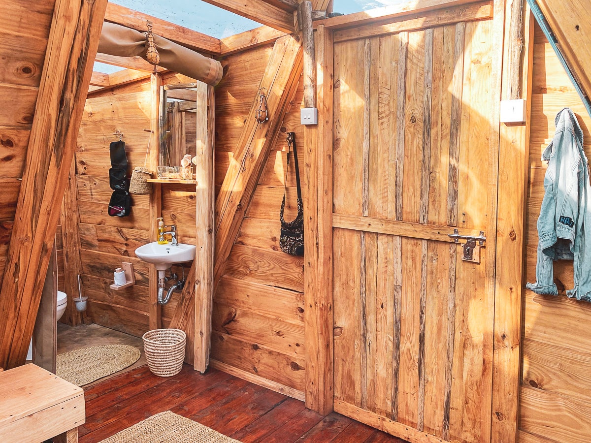 The interior showcases natural wooden walls and floors, providing a cozy, rustic feel. A small washbasin with toiletries is seen alongside a mirror. Various hooks are mounted for hanging essentials, while a woven basket rests on the floor. Natural light filters through an overhead window.