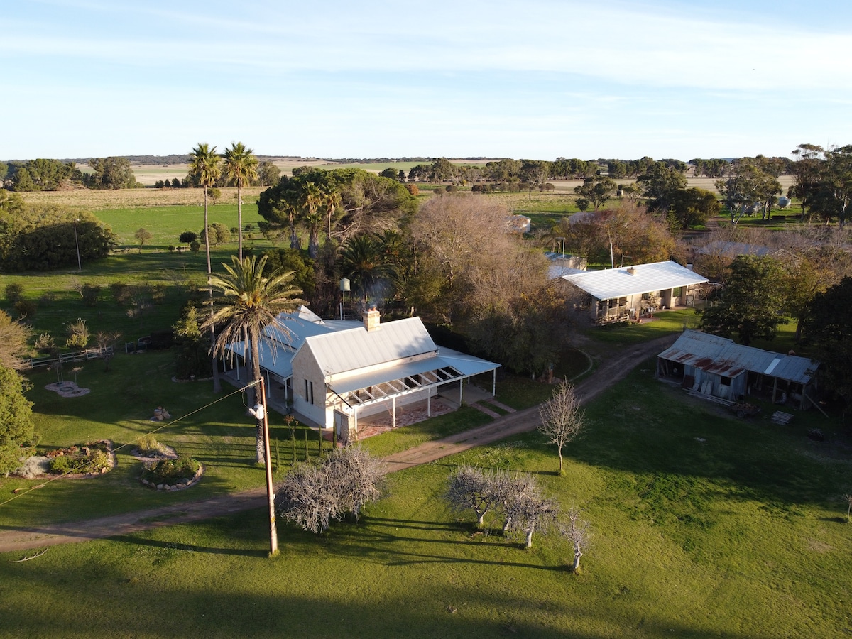 An aerial view showcases the Poona Homestead Cottage and its surrounding grounds, featuring lush green lawns, trees, and gardens. The cottage, with a metal roof, is complemented by a nearby outbuilding. The landscape reflects a tranquil countryside setting.