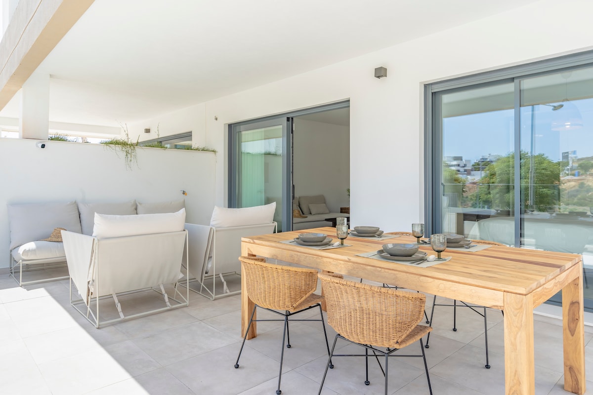 An outdoor terrace features a wooden dining table set for four, accompanied by four woven chairs. Two lounge areas, each with two white armchairs, provide a relaxed seating arrangement. Sliding glass doors lead into the interior spaces, allowing for a seamless transition.