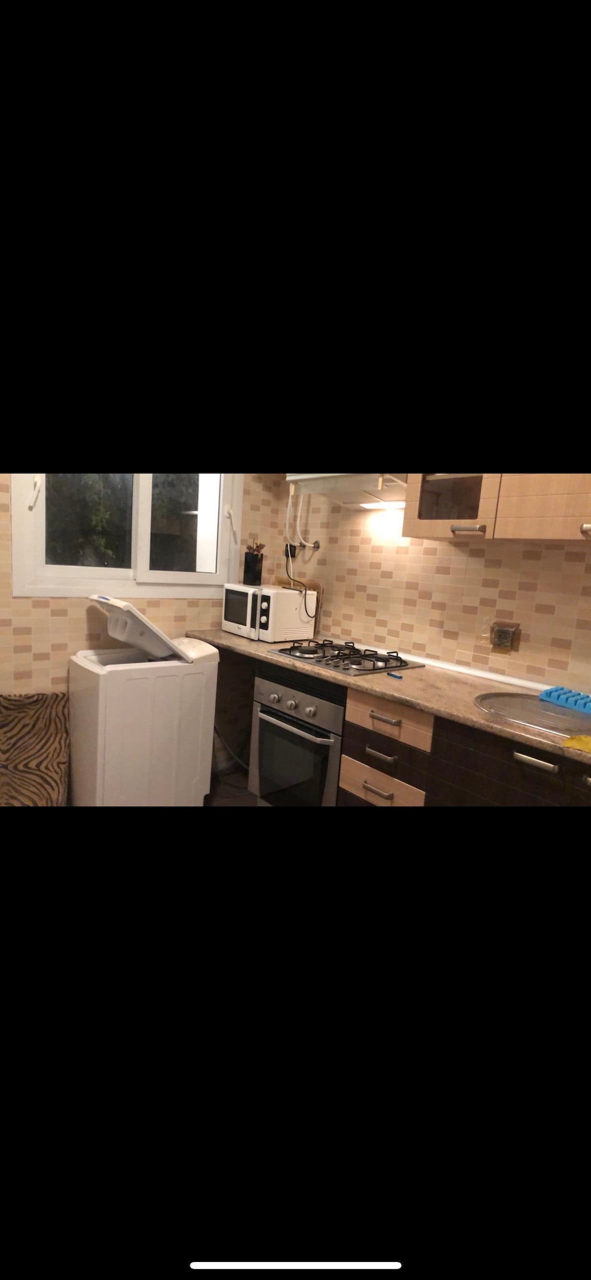 A compact kitchen displays a washing machine alongside a microwave and stove. Light-colored cabinets complement the neutral-toned backsplash. A sink is situated beneath a window, allowing for natural light to illuminate the work area.