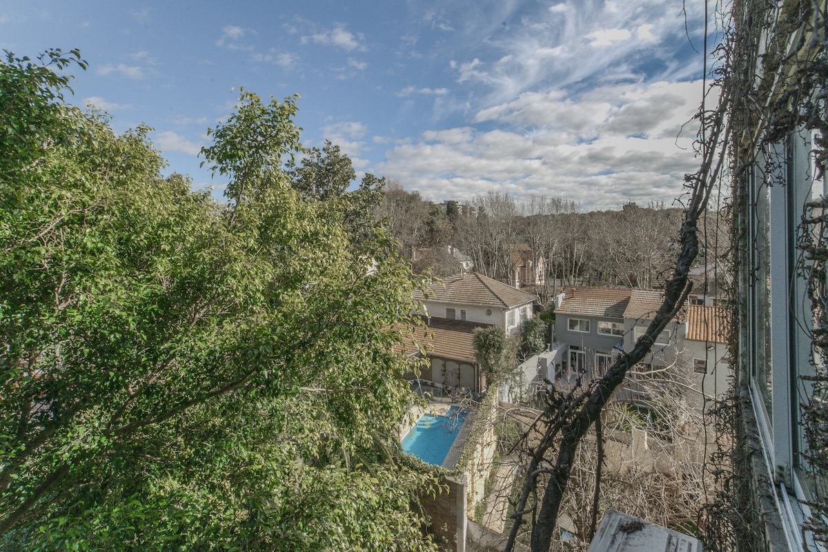 A view of a quiet neighborhood is presented, featuring low-rise houses and green trees. A glimpse of a pool is visible among the residences. The sky exhibits a mix of clouds and blue patches, adding to the serene atmosphere of the area.