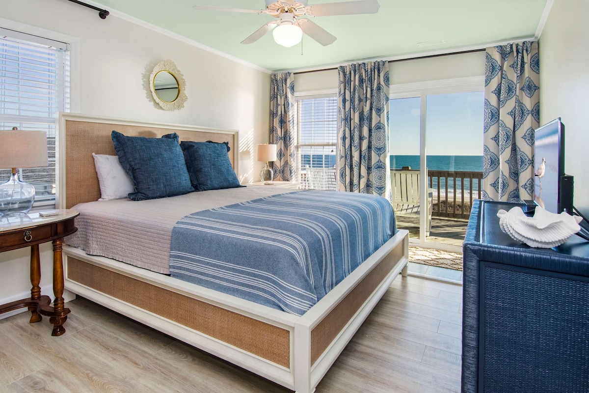 A bright bedroom features a spacious bed with blue and white striped bedding, complemented by large pillows. Floor-to-ceiling windows offer ocean views and natural light. Light-colored walls are accented by soft curtains, while a small bedside table holds lamps, adding functional elegance to the space.