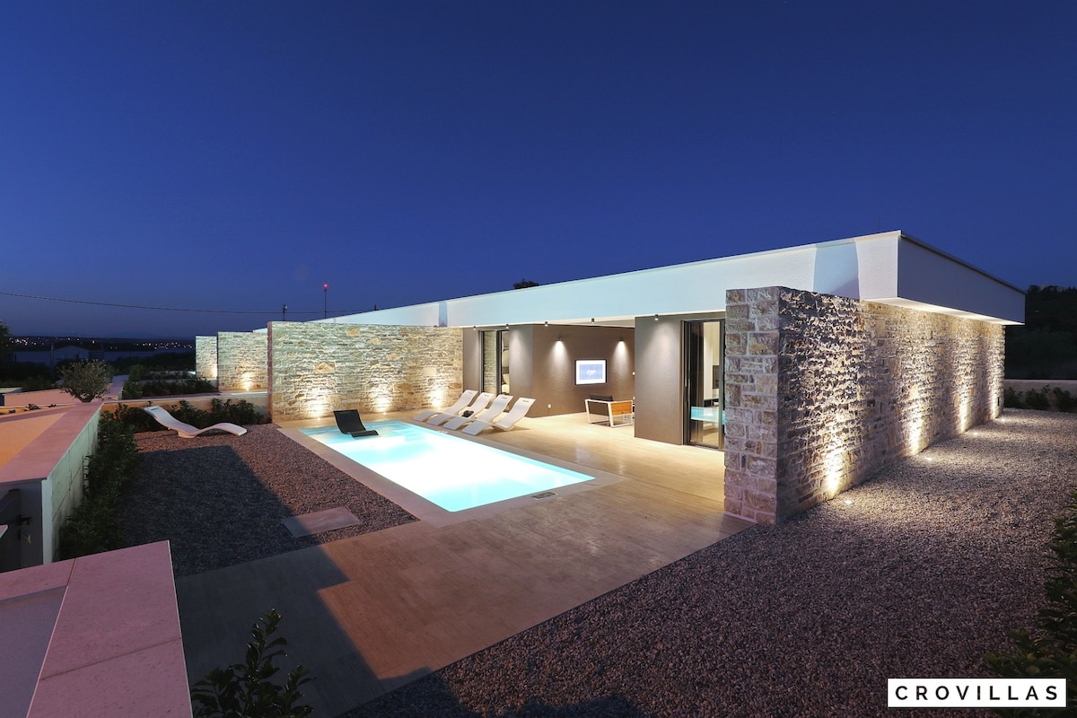 A modern villa is illuminated at dusk, featuring a serene swimming pool surrounded by a textured stone wall. Lounge chairs are arranged for relaxation. The smooth patio extends around the pool, with landscaped gravel highlighting the architecture.