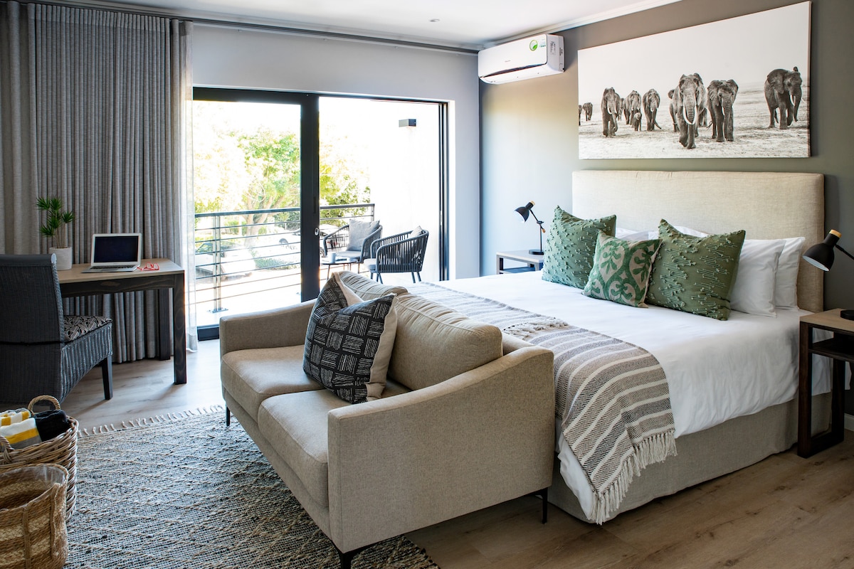 A bright room features a king-sized bed adorned with decorative pillows and a light quilt. A comfortable couch sits in front, while a workspace with a laptop is present. Large glass doors open to a balcony, with artwork depicting elephants on the wall.