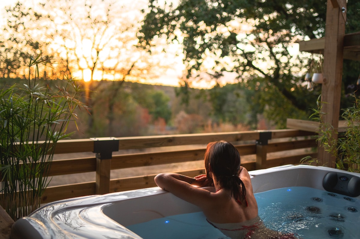 A jacuzzi is set on a wooden terrace, surrounded by natural greenery. The sunset casts a warm glow over the landscape, while a person relaxes in the tub, facing the horizon. The scene offers a sense of tranquility in an intimate outdoor setting.