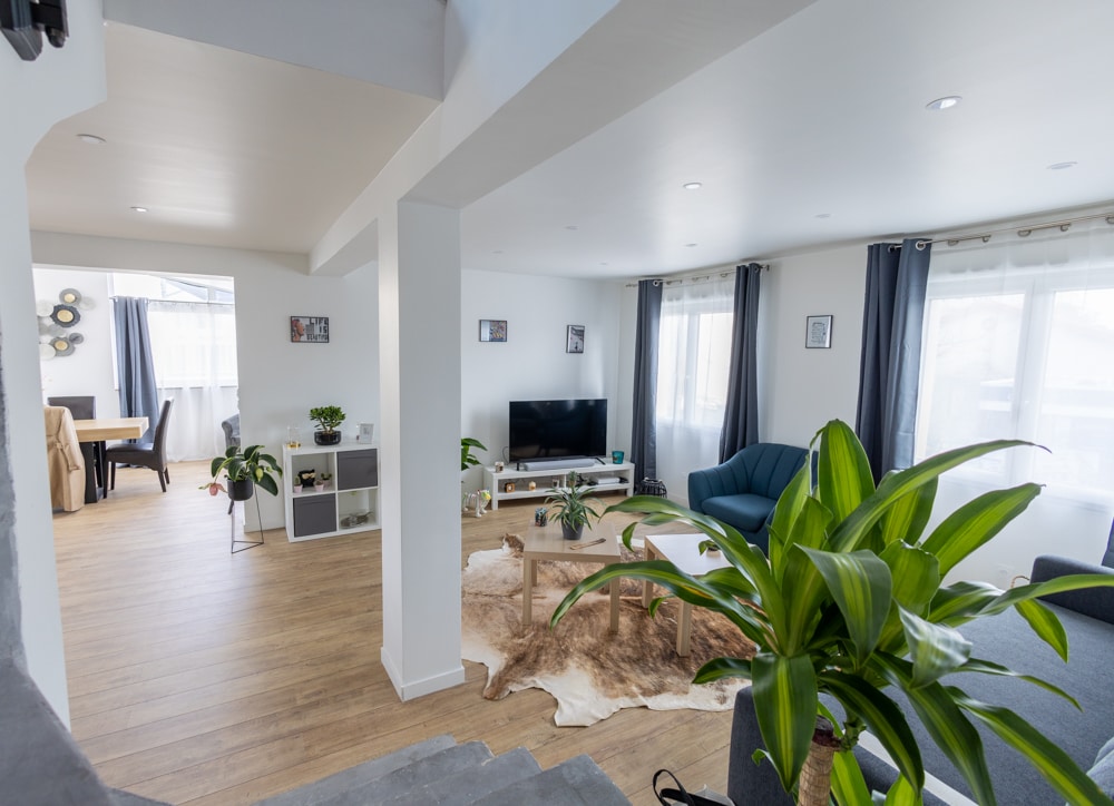 A spacious living area features large windows allowing natural light to fill the room. Comfortable seating is arranged around a wooden coffee table on a textured rug. Potted plants add a touch of greenery, while light-colored walls contribute to a bright and open atmosphere.