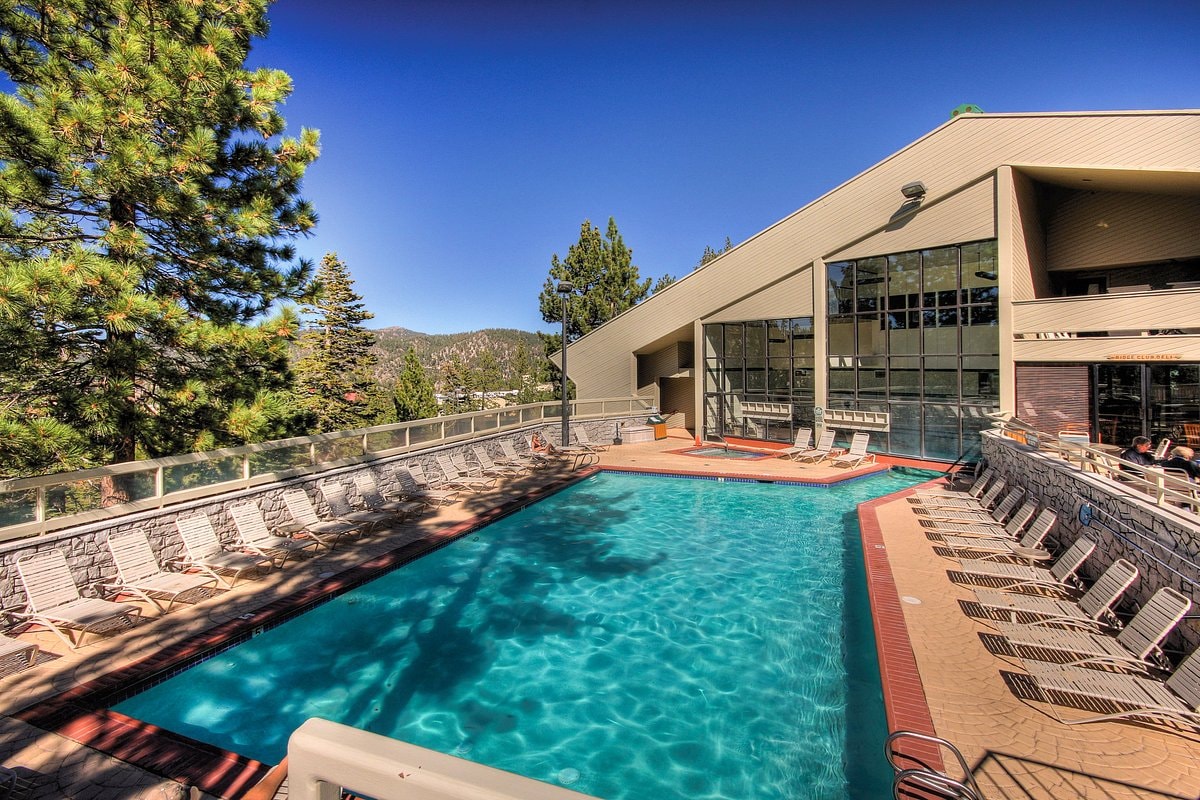 An outdoor heated pool is surrounded by a spacious deck furnished with lounge chairs. The pool is framed by tall trees and offers scenic mountain views. Large windows of the adjacent building let in natural light, creating a bright and inviting space.