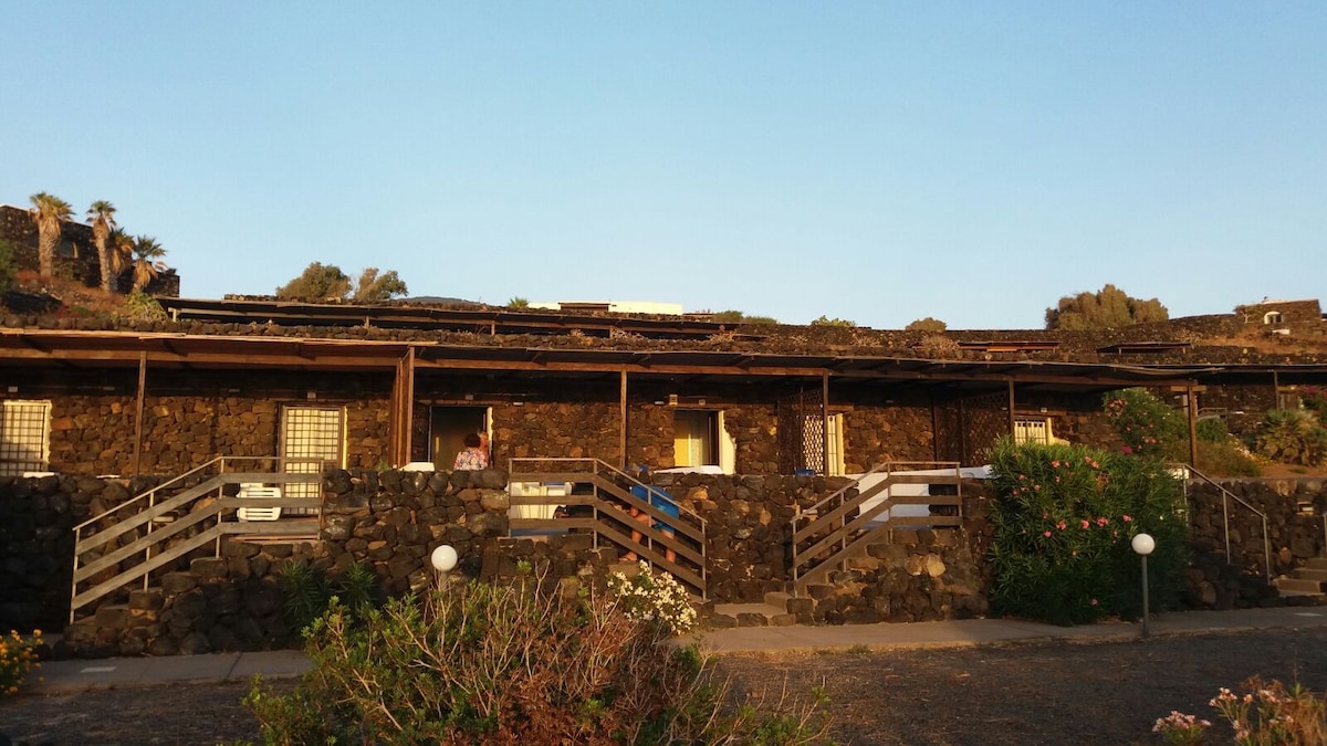 A stone building is viewed under a clear sky, featuring a row of windows and wooden balconies. Softly lit gardens with flowering plants and pathways lead to the structure, enhancing the natural surroundings.