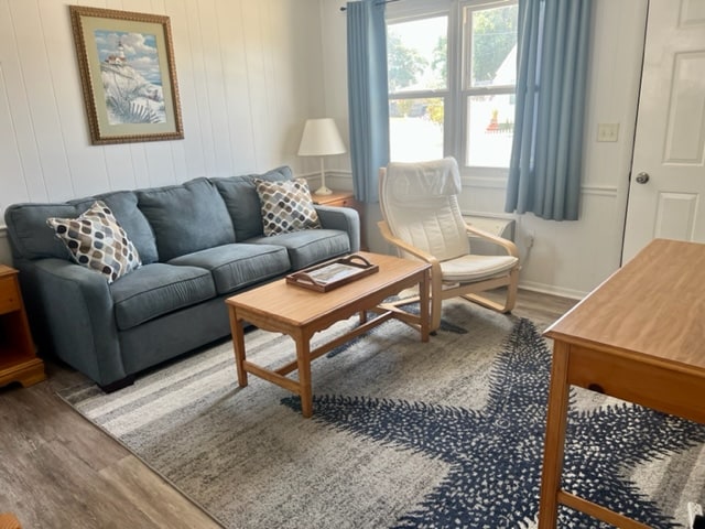 A living area is presented with a blue sofa adorned with patterned pillows, positioned beside a light wood rocking chair. A wooden coffee table sits in front, accompanied by a simple decorative tray. Natural light enters through two windows dressed with light blue curtains.