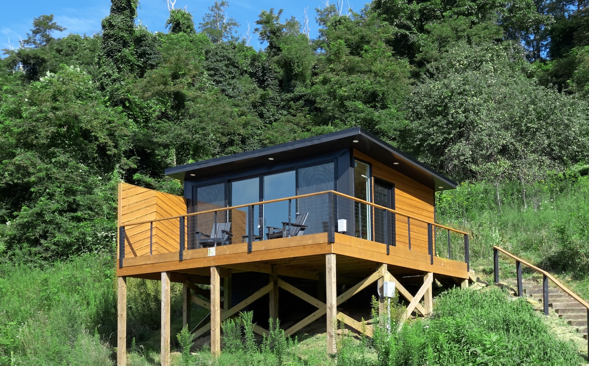 A modern glass cabin is elevated on wooden stilts, surrounded by lush green foliage. Large panoramic windows frame the outdoor deck, which features seating for relaxation. The structure's blend of natural wood and sleek design harmonizes with its mountain setting.