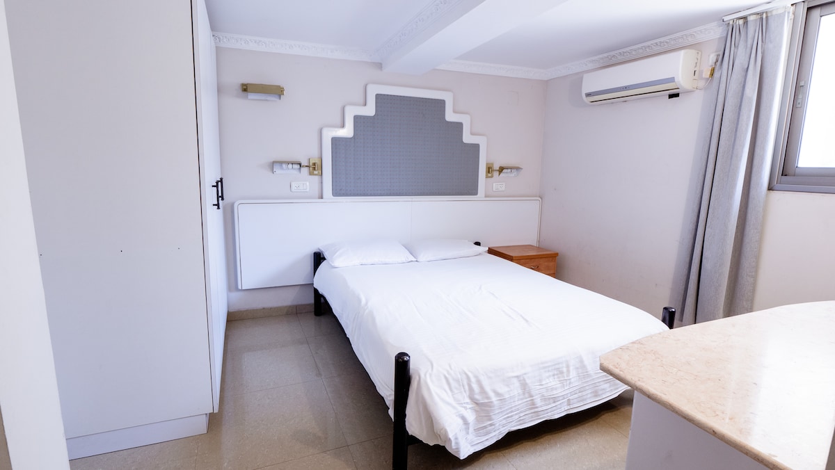 A simple sleeping area is presented with a spacious bed covered in white linens. A wood nightstand is positioned beside the bed. Natural light enters through a window, complemented by light grey curtains. An air conditioning unit is mounted on the wall for comfort.