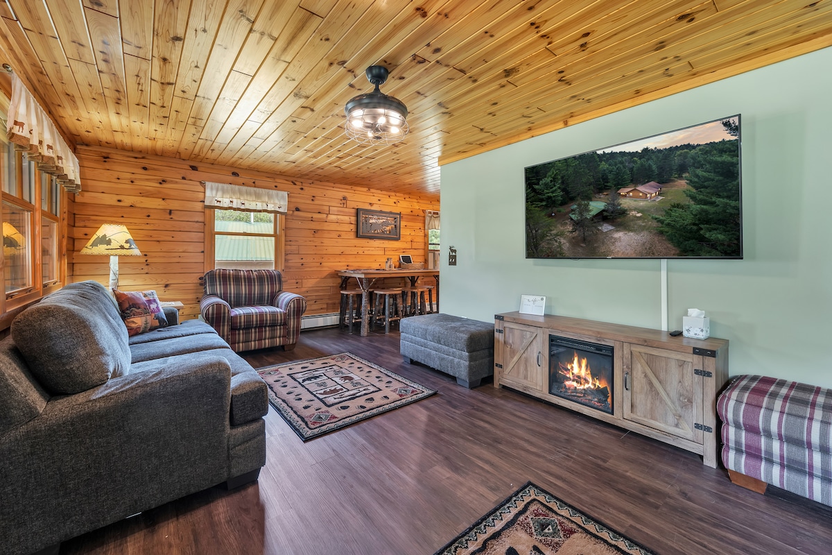 A welcoming living area features a combination of dark sofas and plaid chairs. A large flat-screen TV is mounted on a wooden wall, displaying outdoor scenery. A cozy fireplace sits below the TV, providing a warm ambiance. Wooden paneling and ceiling enhance the natural feel.