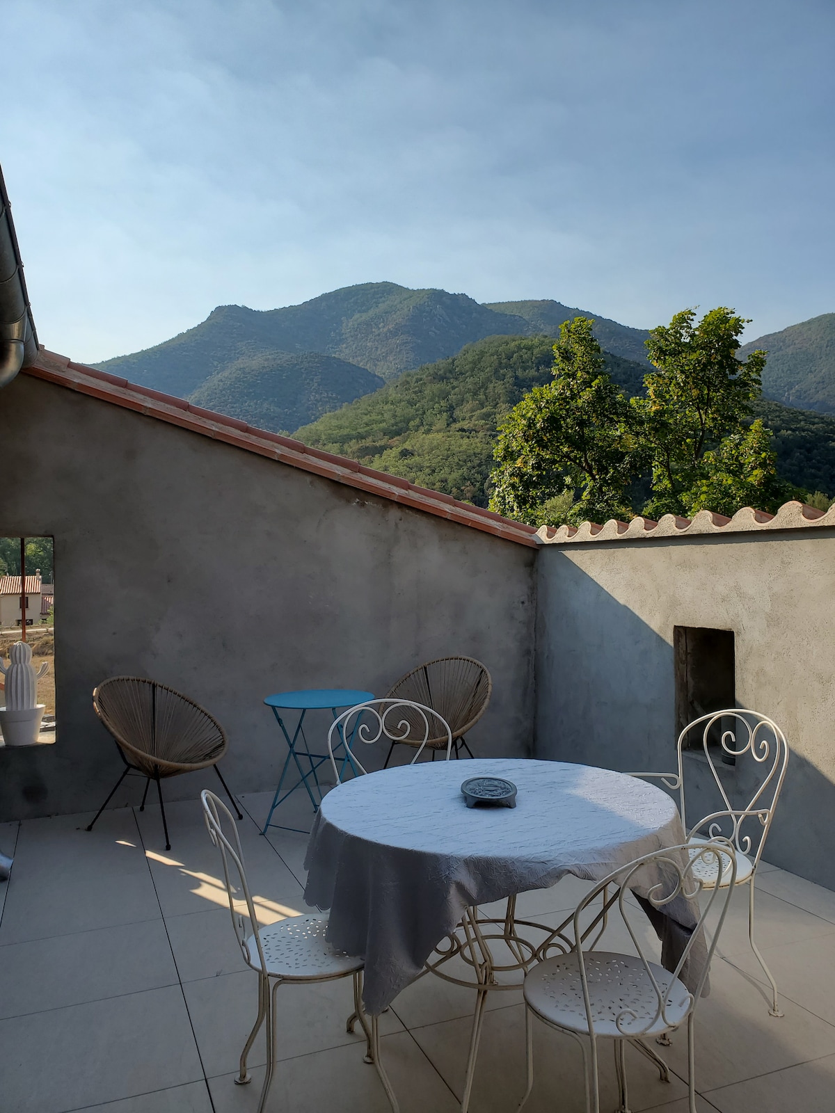 A scenic terrace features a round table with four chairs, surrounded by mountains in the background. Two Acapulco-style armchairs add comfort, while a small blue table provides additional seating. Natural light illuminates the space, enhancing the tranquil atmosphere.