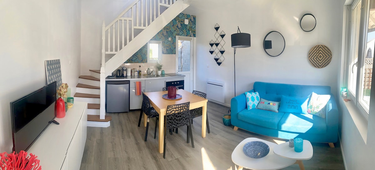 The open living area features a bright blue couch and a light wood dining table surrounded by black chairs. Natural light fills the space through large windows, highlighting a modern kitchen area with stainless steel appliances. Decorative items add character to the walls.