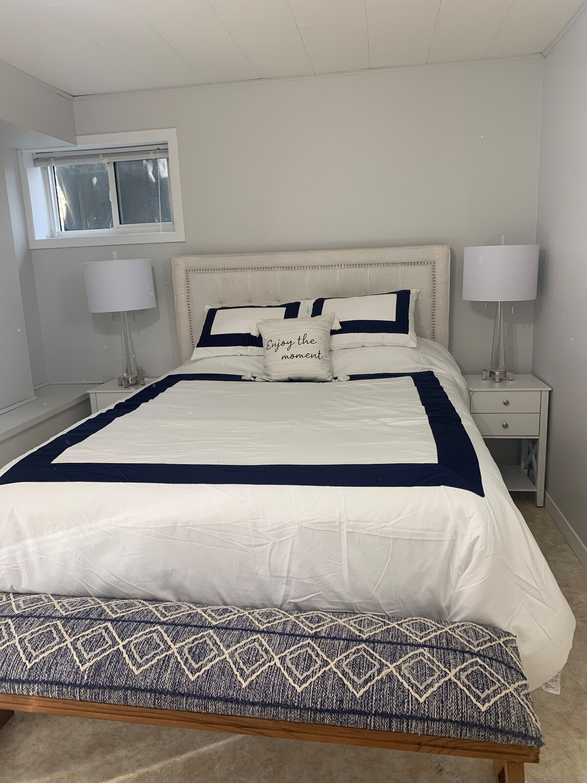 A well-lit bedroom features a spacious bed with a neatly arranged white and navy bedding set. Two bedside lamps provide illumination on either side of the bed. A decorative bench is placed at the foot of the bed, with natural light streaming through a nearby window.