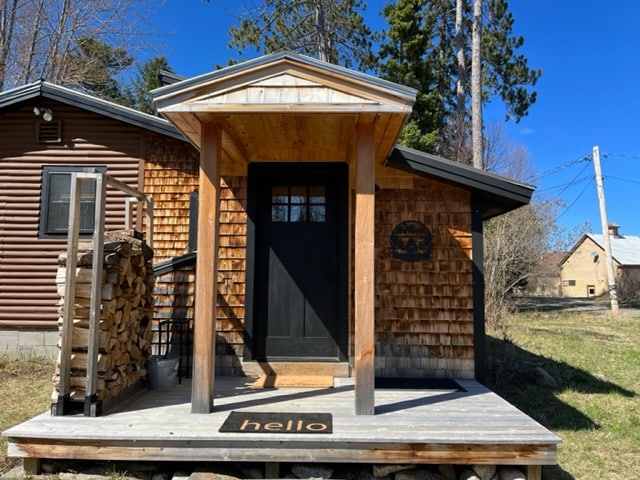 The entrance features a wooden porch with a welcome mat at the front door. A rustic wooden canopy provides shelter, while the exterior showcases natural wood shingles complemented by a dark door. Sunlight illuminates the clear blue sky above.