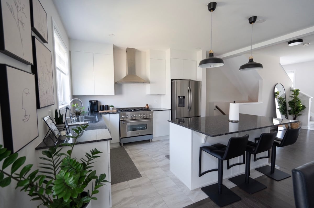 The kitchen features modern stainless steel appliances and a spacious island with seating for three. White cabinetry and granite countertops create a bright workspace, while a large refrigerator and coffee maker provide convenience. Artwork and greenery add subtle touches to the contemporary design.