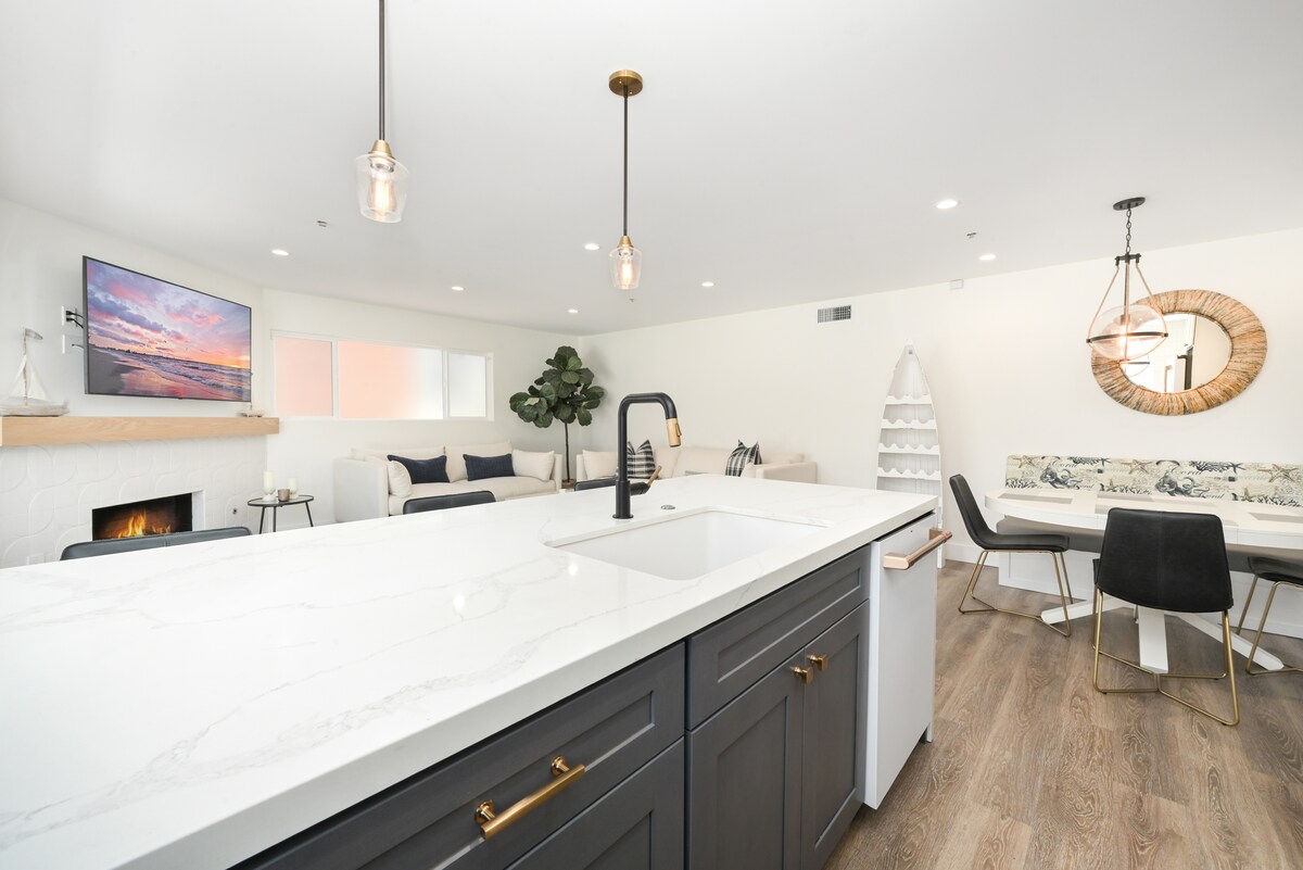 An open-concept living space is depicted, showcasing a large kitchen island with a farmhouse-style sink. A modern television is mounted on the wall, above a white fireplace. A dining area with dark chairs surrounds a table, while a cozy seating area features light-colored sofas and a decorative wall mirror.
