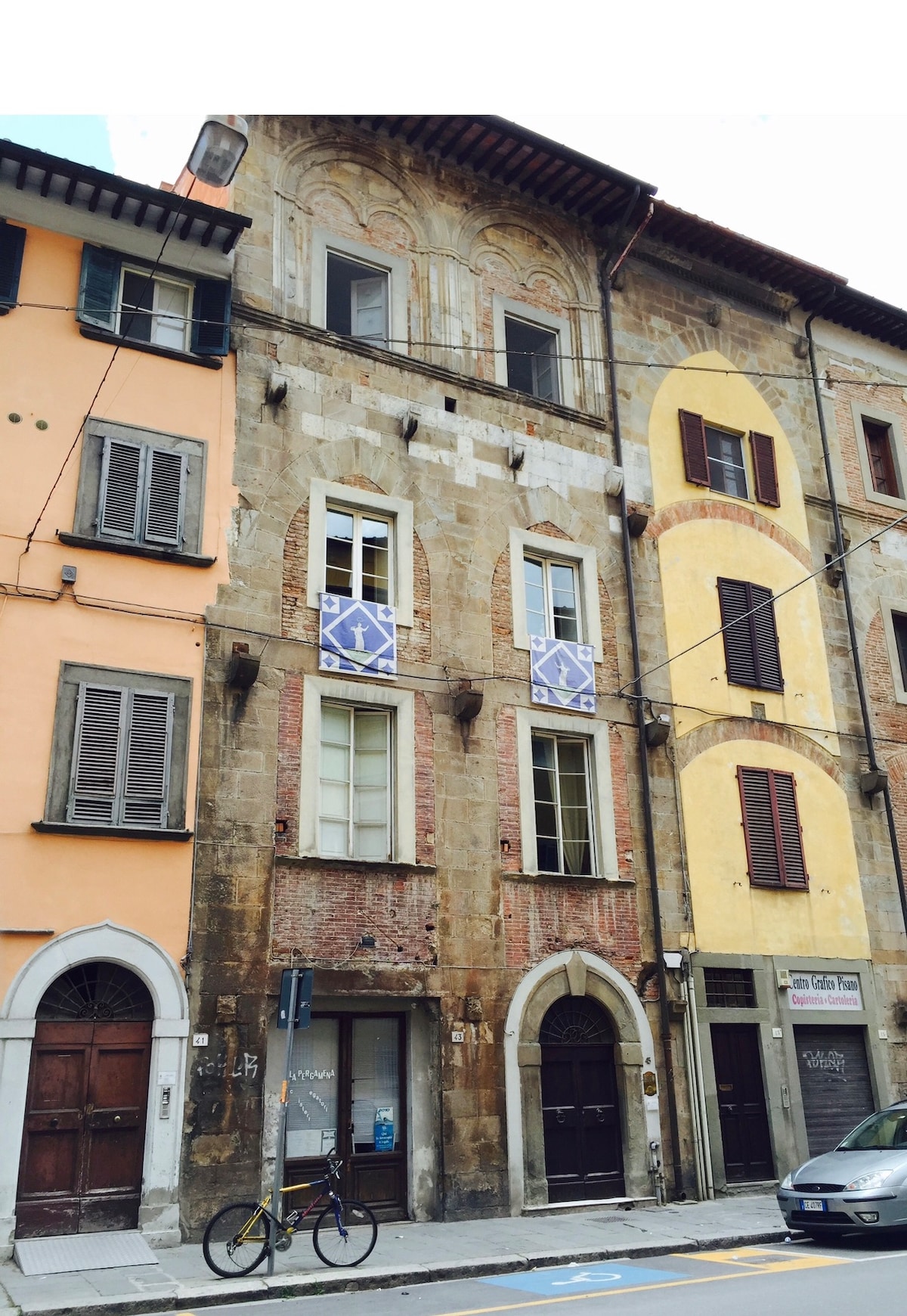 The medieval tower house features a distinctive façade with a combination of stone and brick, showcasing ornate window detailing. Two blue signs with directional arrows are attached to the exterior. Different colored buildings flank the structure, contributing to the historic town center atmosphere.
