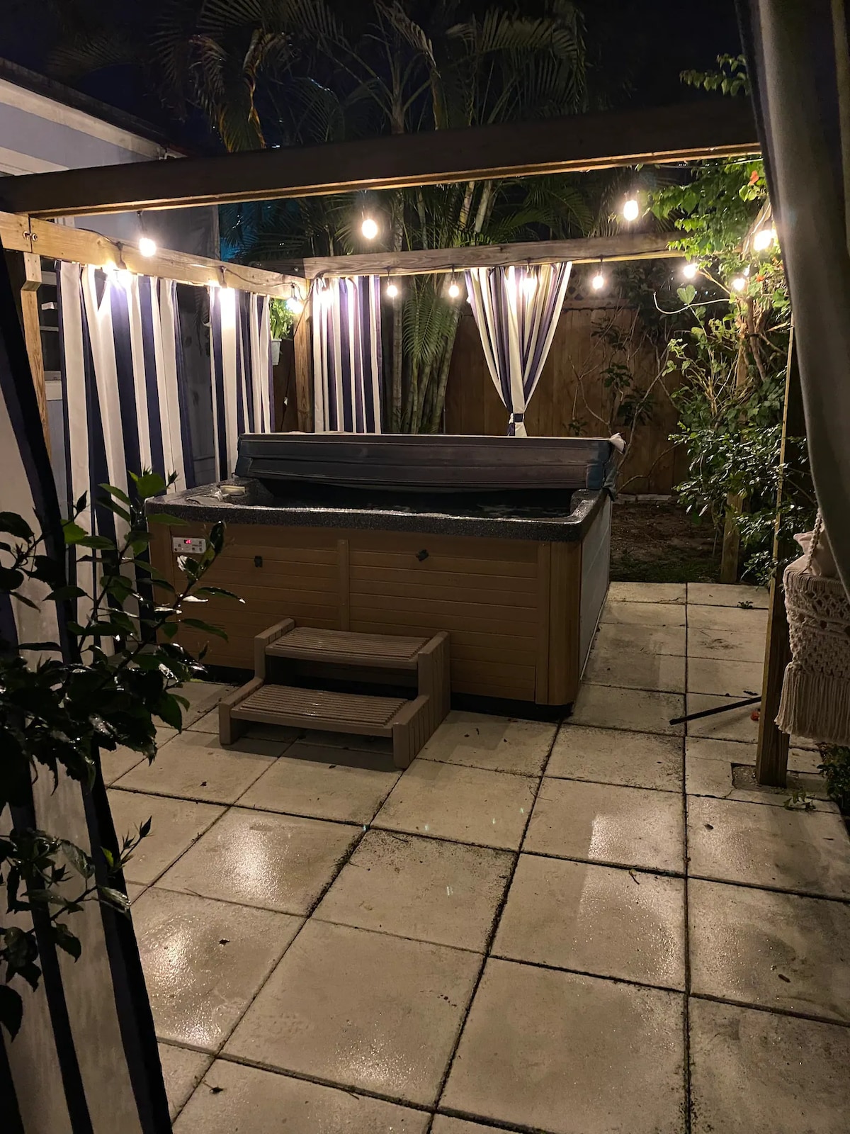 A shared hot tub is situated under a wooden pergola adorned with soft lighting. Curtains drape gracefully, creating a cozy ambiance, while the stone patio provides a harmonious setting surrounded by lush greenery.