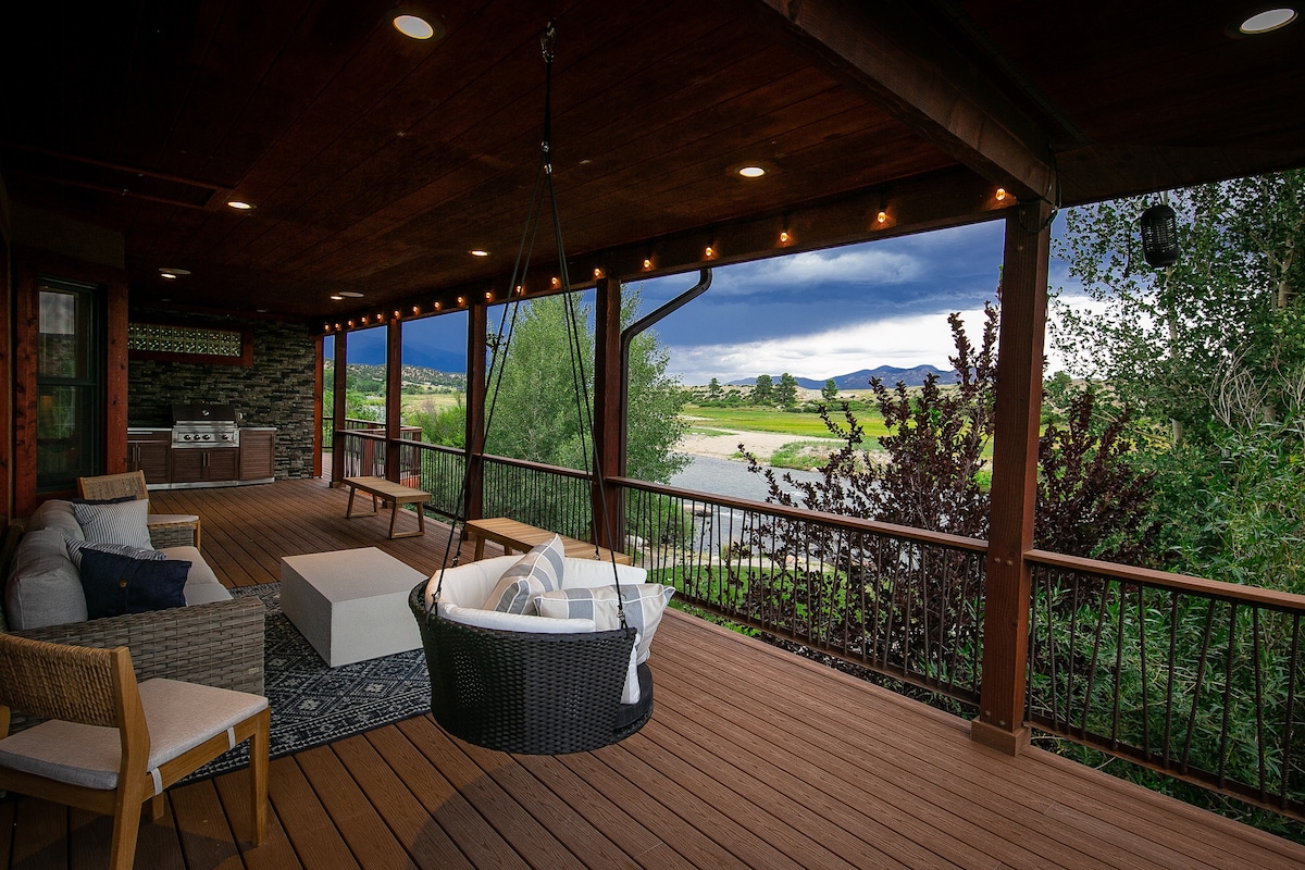 An outdoor deck features a hanging chair and cozy seating area, arranged for relaxing. Soft lighting enhances the space, while views of the Arkansas River and surrounding greenery create a serene backdrop. A gas grill is visible in the corner, inviting outdoor dining.