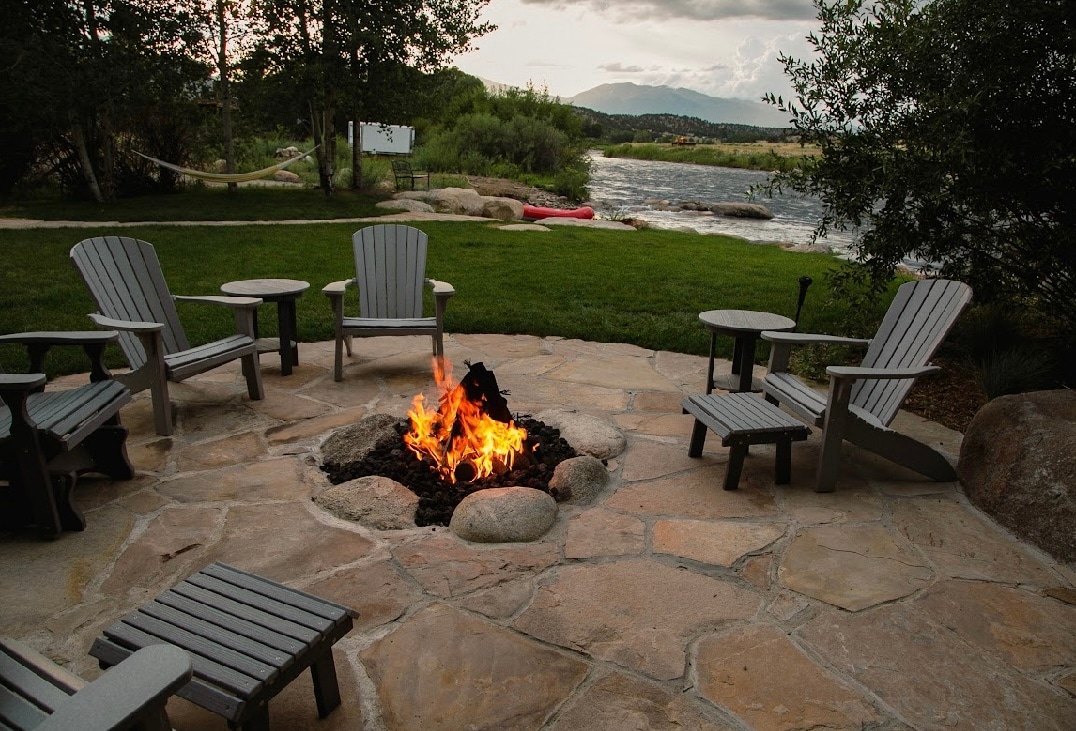 An outdoor fire pit surrounded by several wooden chairs is situated on a stone patio. The calm Arkansas River flows nearby, with a backdrop of lush greenery and mountains. A hammock can be seen in the background, providing a relaxing space for guests.