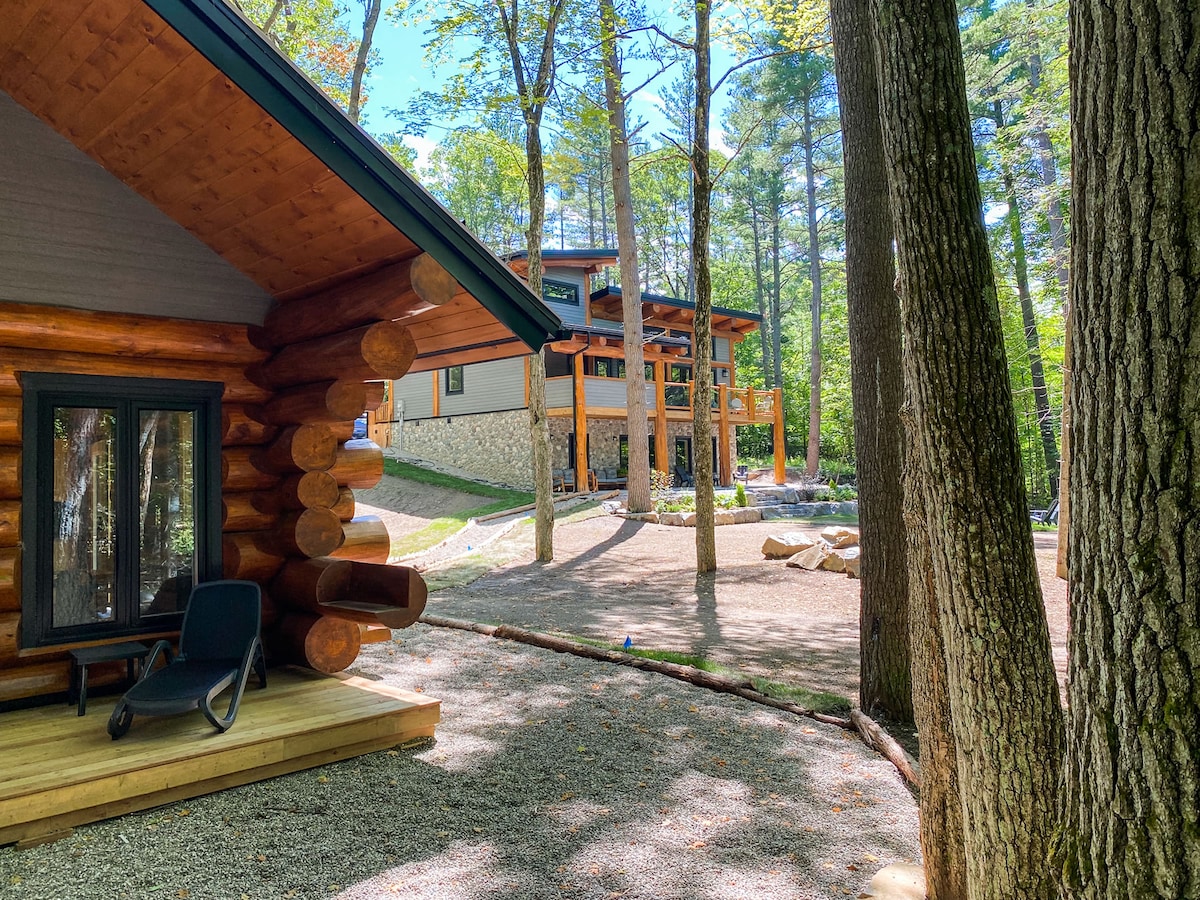 A cozy log structure is set against a backdrop of tall trees, with a well-maintained gravel path leading to a spacious main cabin. A simple chair sits on the deck, inviting relaxation in a peaceful outdoor atmosphere.