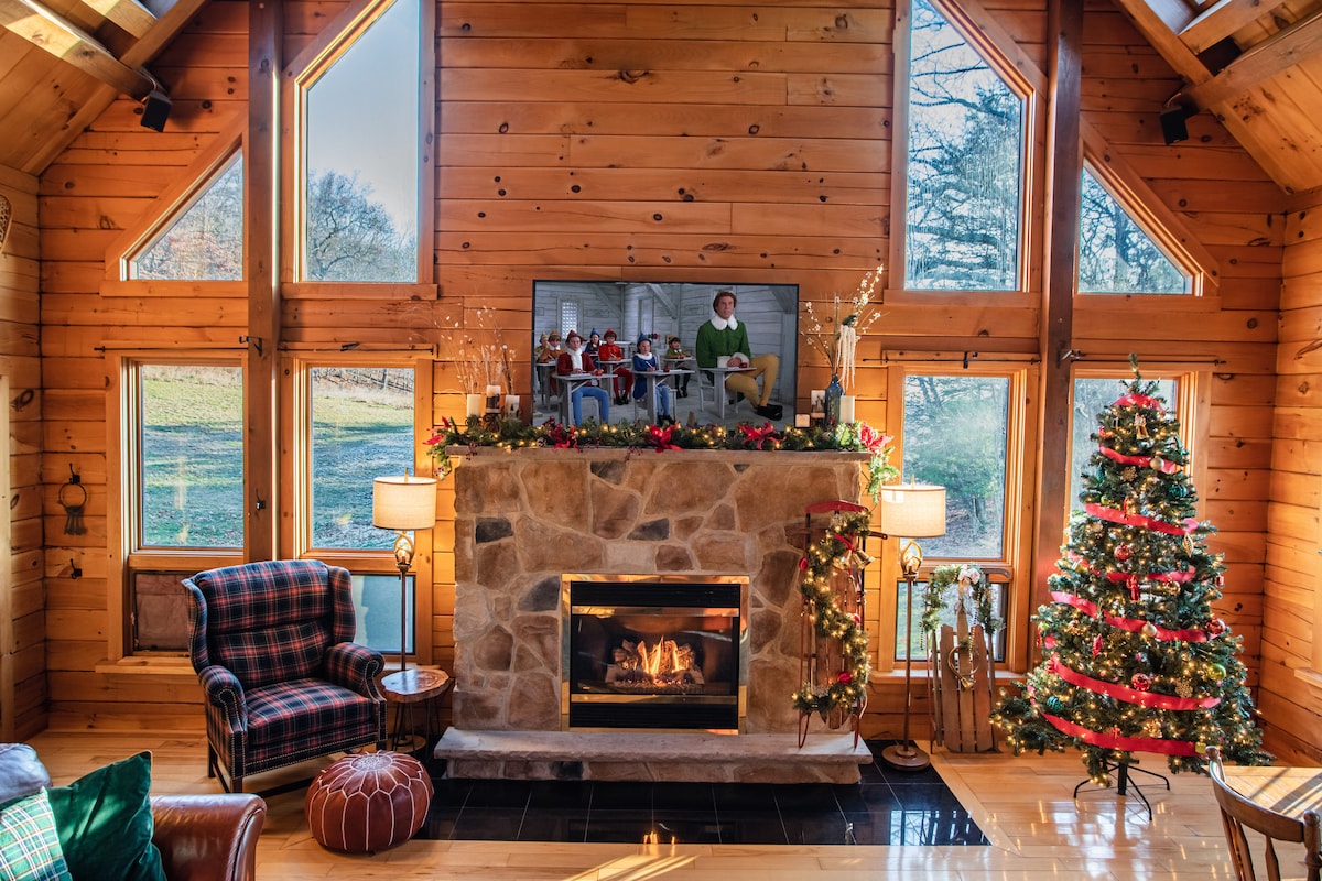 A cozy living area is highlighted by a stone fireplace with a television above, all framed by large windows allowing natural light. A Christmas tree adorned with decorations stands nearby, complemented by two armchairs and soft lighting providing warmth to the space.