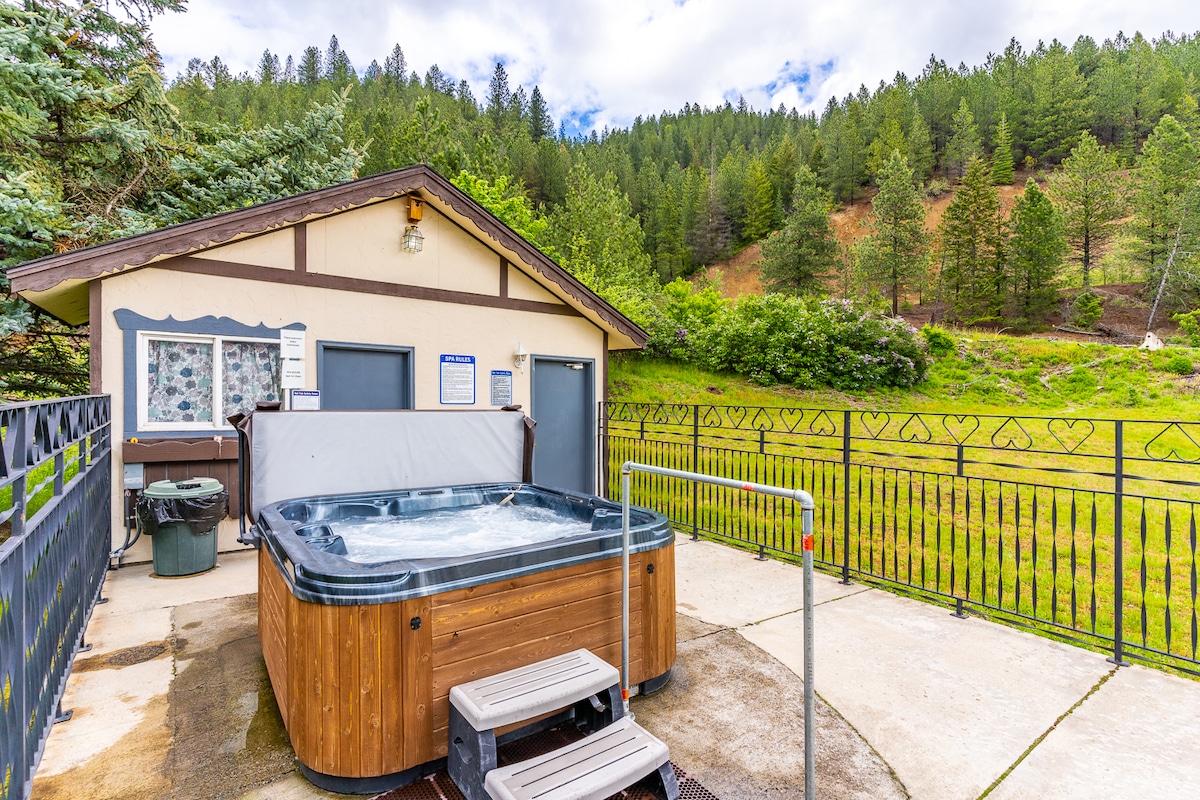 A community hot tub is situated on a concrete patio surrounded by a gated area. Lush greenery and trees are visible in the background, providing a serene atmosphere. The hot tub is accompanied by steps for easy access.