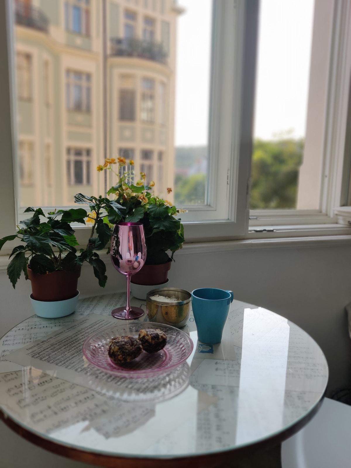 A small table is set next to a large window, featuring a pink glass filled with a beverage and a blue cup. A plate with two baked goods rests on the table. Potted plants and yellow flowers bring a touch of greenery to the scene, contrasting with the architectural view outside.