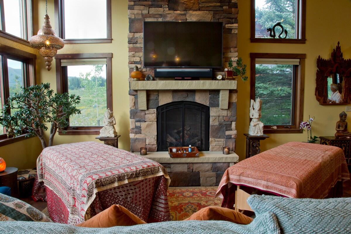 A cozy living area is highlighted by a stone fireplace framed by large windows, allowing natural light and views of the surrounding landscape. Two massage tables are positioned comfortably, draped with vibrant textiles, adding a calming touch to the space decorated with art and plants.