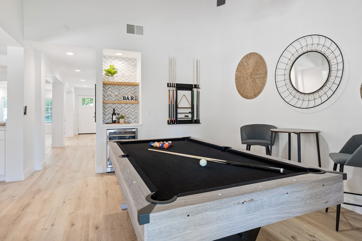 An open game area displays a sleek billiard table with a dark cloth and colorful balls arranged for play. Stylish seating is positioned nearby, complemented by a wall-mounted bar area, circular mirror, and decorative wall accents, enhancing the inviting space.