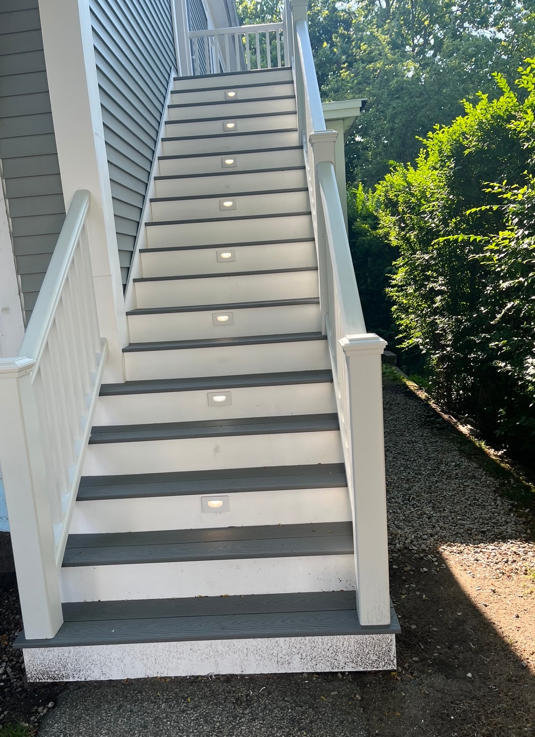 The well-lit staircase features alternating lights embedded in the steps, providing visibility and guidance. The light grey stairs contrast with the white railings, leading up to a welcoming entrance surrounded by greenery. The pathway beside the stairs is lined with gravel.