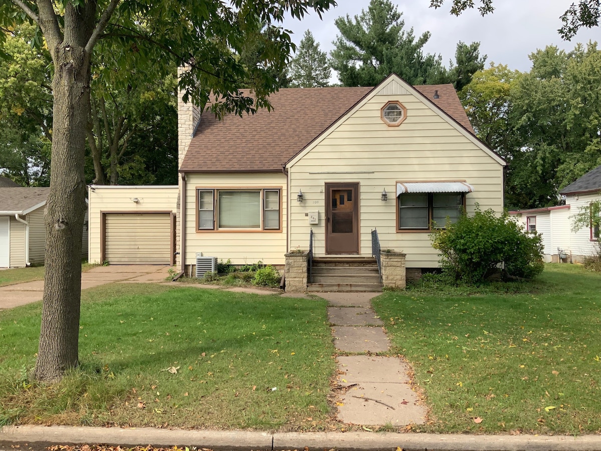 The exterior of a charming 1950s home is visible, featuring a sloped roof and light-colored siding. A short path leads to the front door, surrounded by a well-kept lawn and shrubs. The garage is attached on the left side, complementing the house's character.