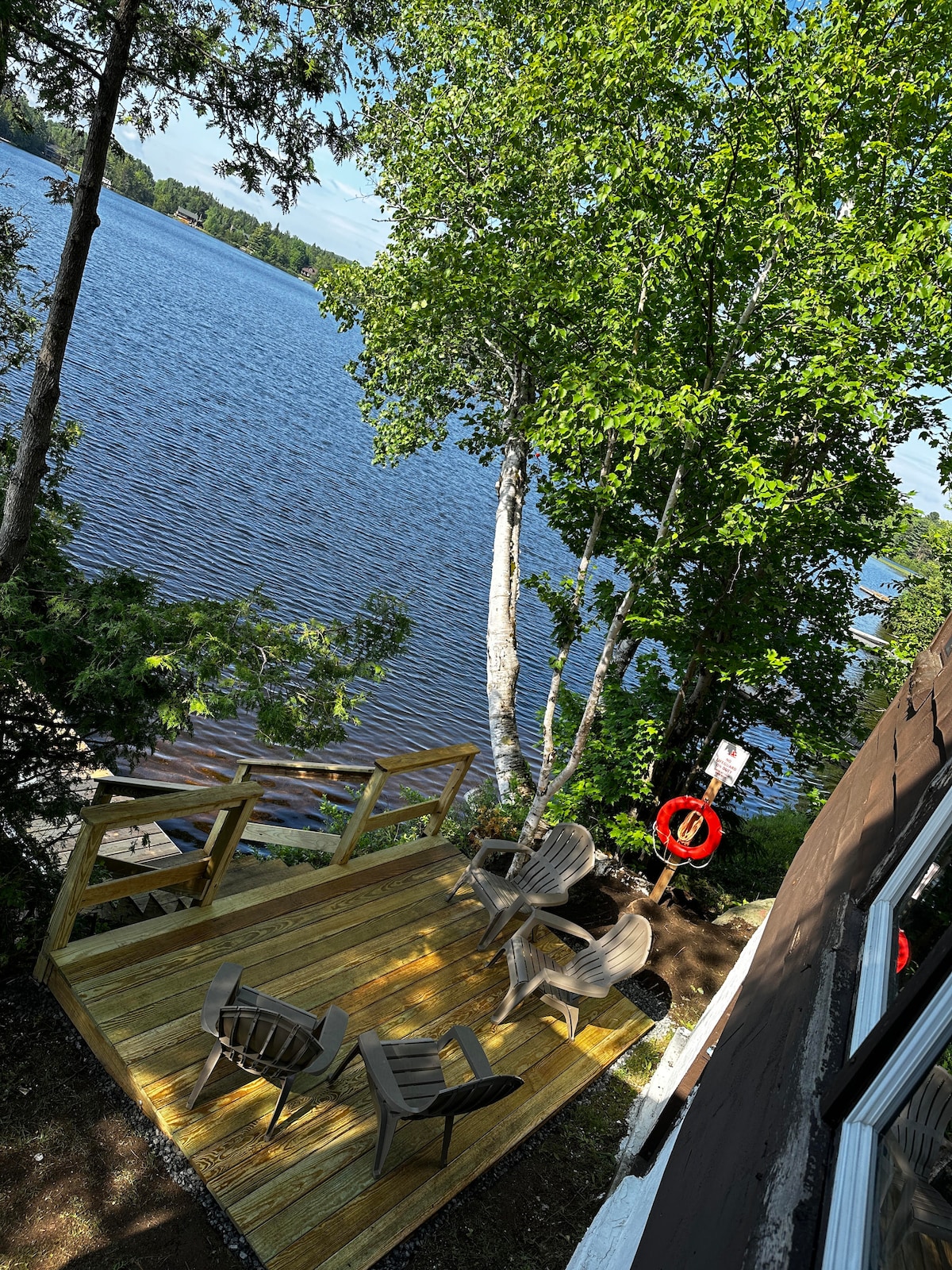 A wooden deck located at the water's edge is furnished with several chairs, inviting relaxation. Trees provide shade, while the lake is visible beyond, with calm waters reflecting the sky. A life ring hangs on a post nearby, enhancing safety and accessibility.