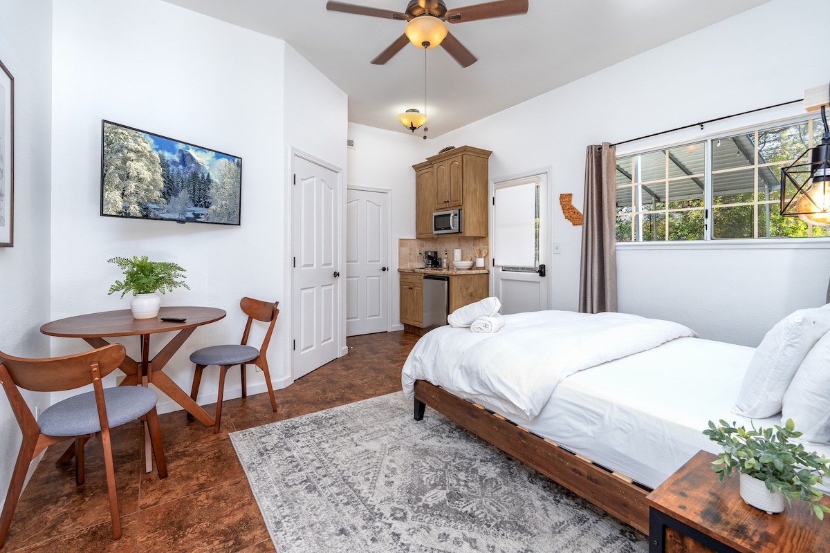 An inviting guest room features a spacious bed with a white comforter. A small dining area with two chairs and a table is situated near a window, providing natural light. A kitchenette equipped with appliances is visible, alongside a mounted TV on the wall.