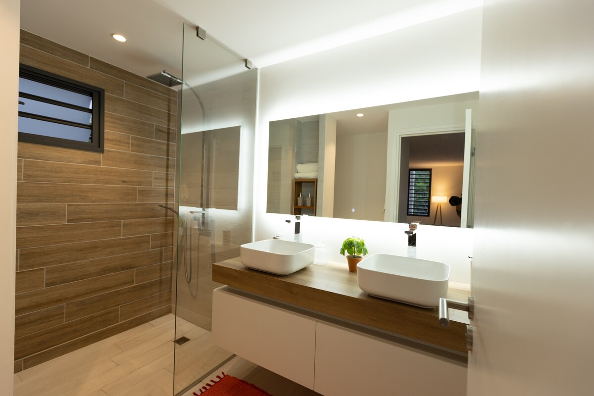 A modern bathroom features a spacious walk-in shower with a glass panel. Two vessel sinks are placed on a sleek countertop below a backlit mirror. Warm wood accents and neutral tiles create a clean, inviting atmosphere, complemented by a small potted plant.