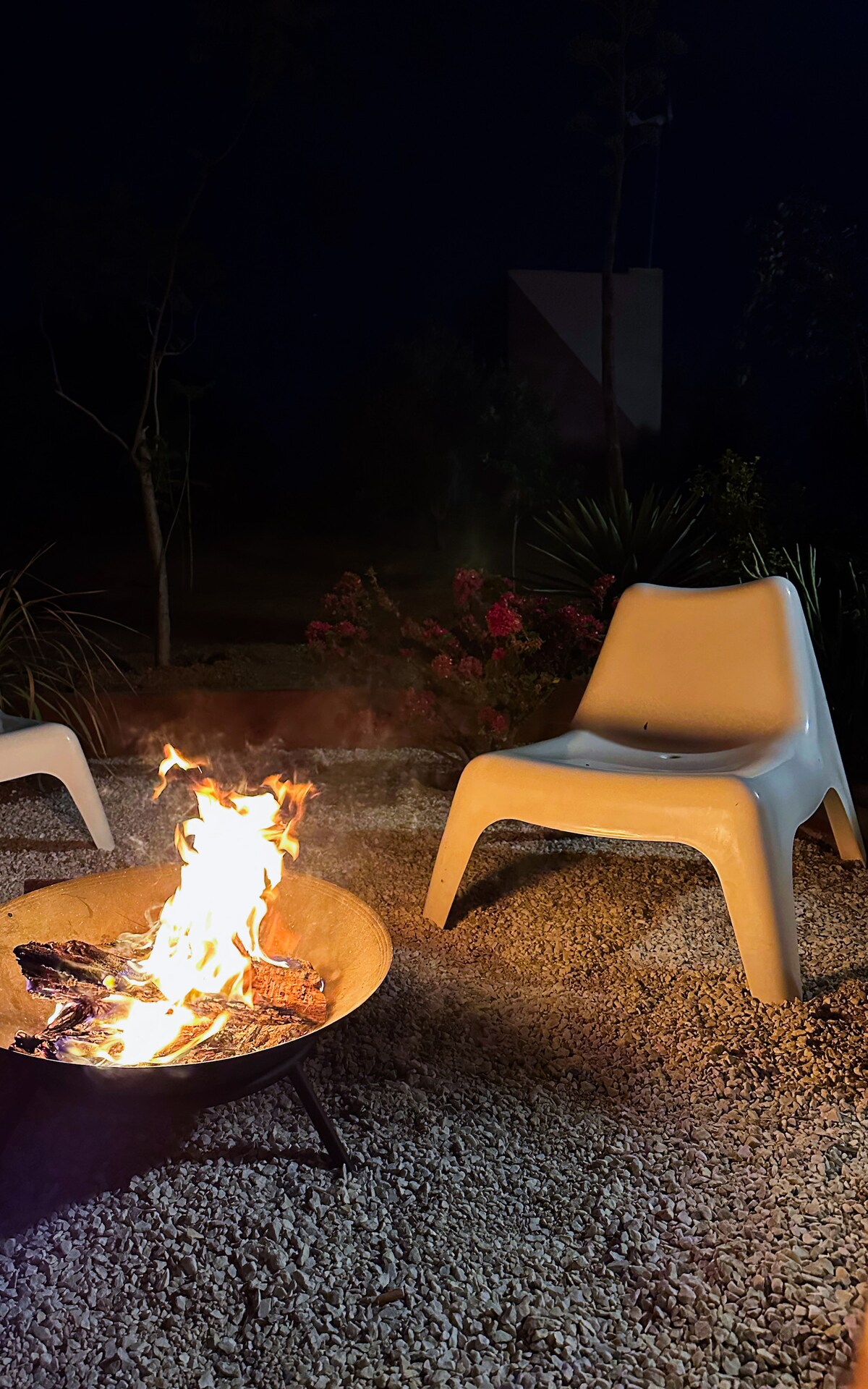 A fire pit is surrounded by a gravel area, with a bright flame illuminating the scene. A simple white chair is positioned nearby, complementing the natural setting. In the background, greenery and flowers are softly visible, enhancing the outdoor atmosphere.