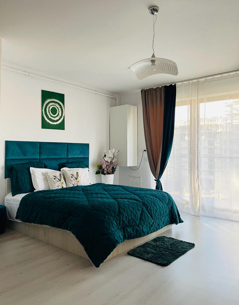 The sleeping area showcases a spacious bed with a dark green quilted coverlet and decorative pillows. A light-colored rug is placed at the foot of the bed. Natural light filters through sheer curtains, creating a bright and airy atmosphere.