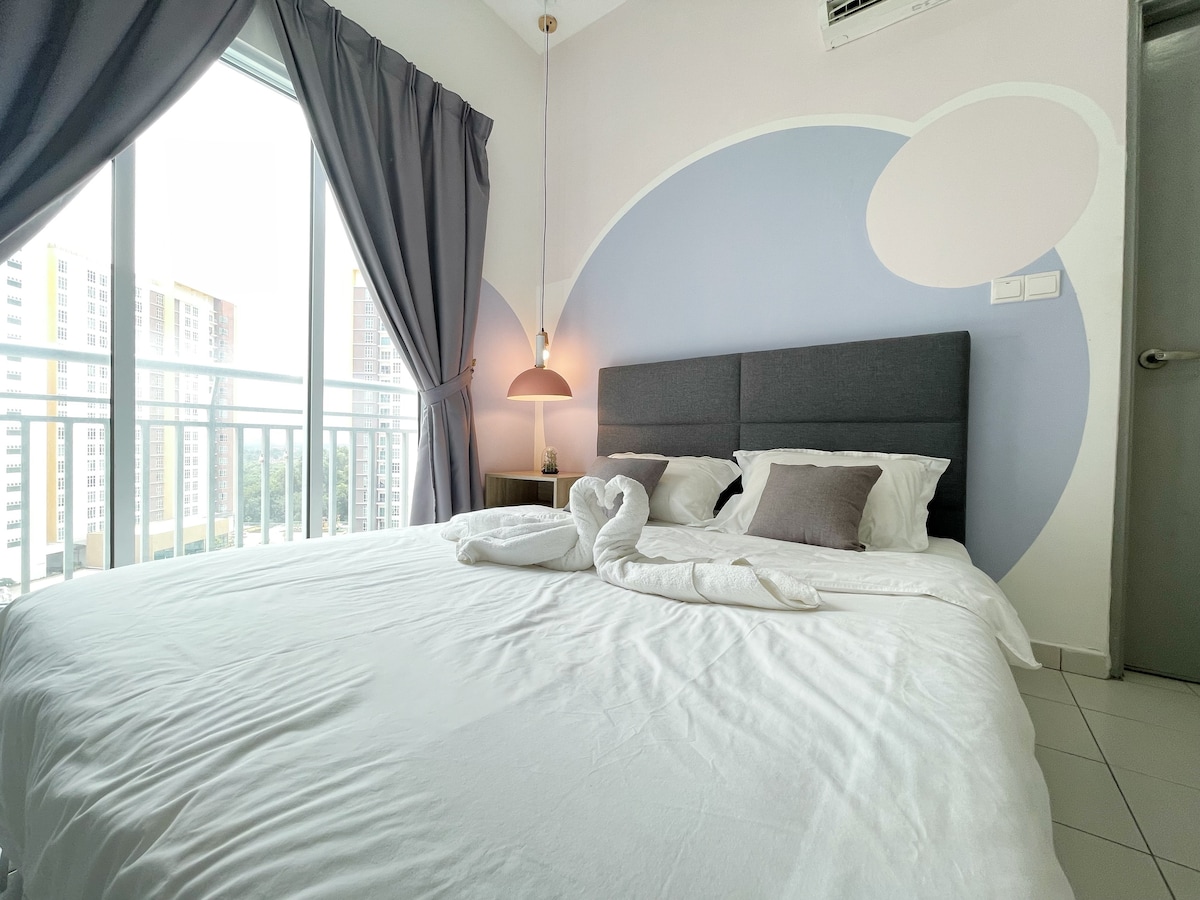 A spacious bedroom features a comfortable bed with crisp white linens and a subtle grey headboard. Soft grey curtains frame large windows, allowing natural light to fill the room. A modern pendant light hangs above a small bedside table, complementing the calming wall design.